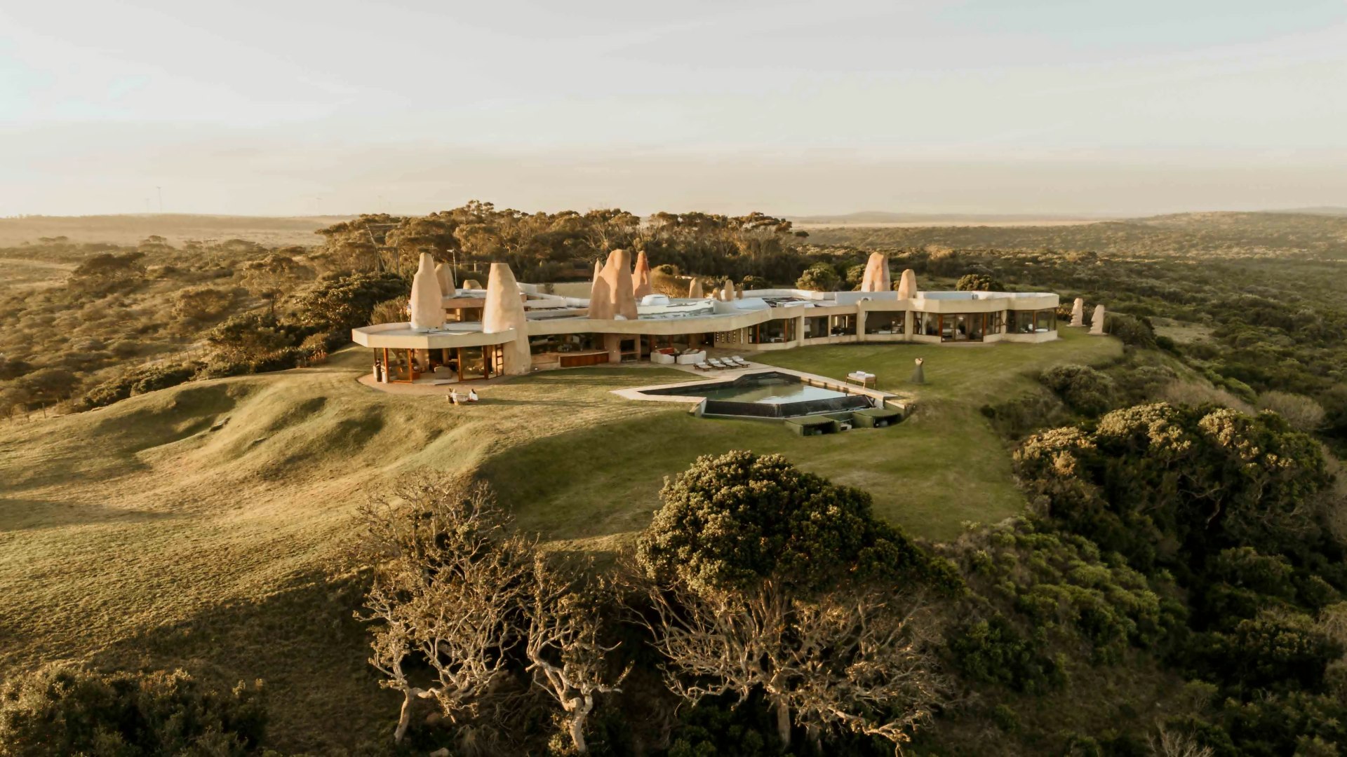 Sandcastle villa photographed from above at golden hour, the infinity pool in front and the reserve hills beyond