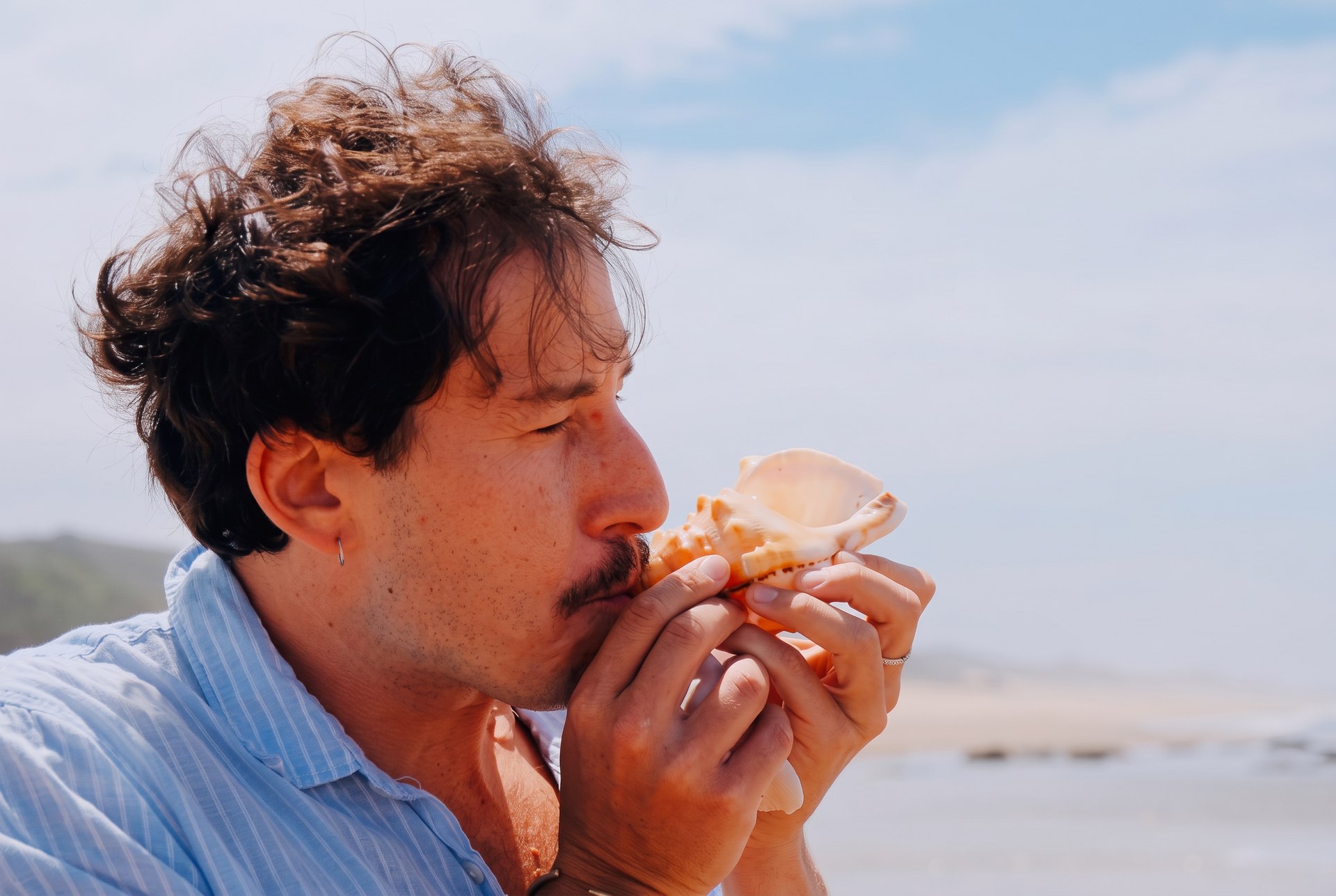 A guest sounding a large conch shell on the beach, the dunes rising behind